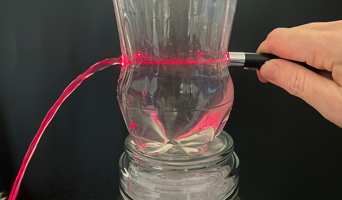 Laser beam shining through a plastic water bottle and continuing insde a stream of water coming out of a hole in the bottle